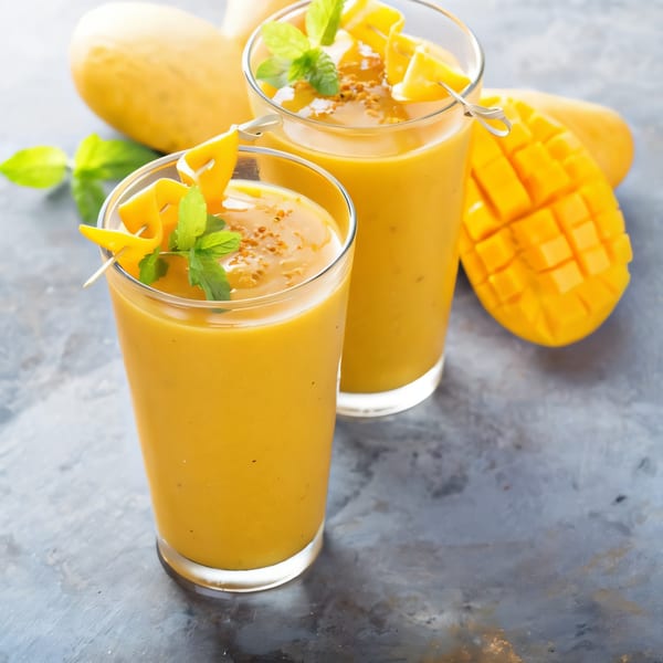 Two glasses of Indian mango lassi, garnished with mint and mango slices, on a gray surface.