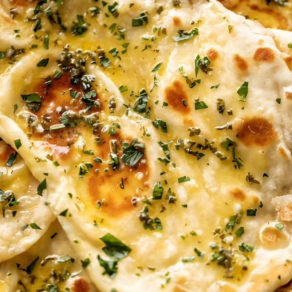 Close-up of golden Indian naan bread, glistening with melted butter and sprinkled with fresh green herbs.