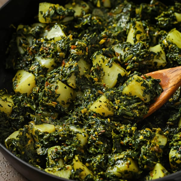 Close-up of Aloo Palak, an Indian dish with potatoes and spinach, in a black pan with a wooden spoon.