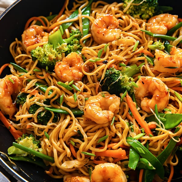 Close-up of shrimp lo mein with noodles, broccoli, carrots, and snow peas in a black pan.